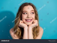 stock-photo-smiling-pretty-young-woman-with-braces-resting-hands-on-face-against-vibrant-blue-background-2651944947